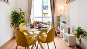 A dining area with yellow chairs, a round table, and a window with curtains. There is a plant on the left and a shelf with books and a vase on the right.