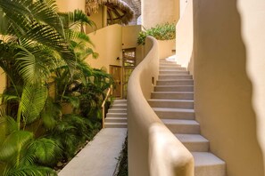 A staircase with a curved handrail leading to a door at the top, surrounded by lush greenery and a thatched roof structure.