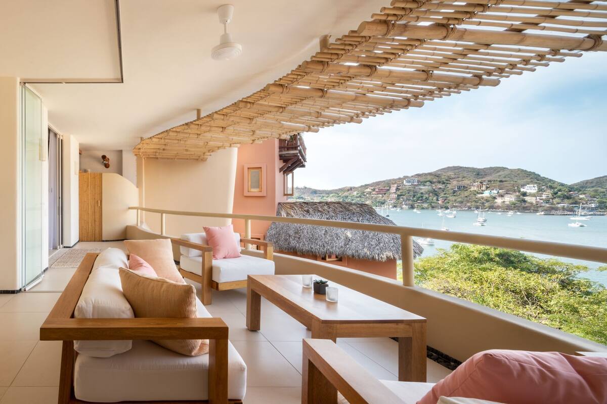 A balcony with a view of the sea, a wooden pergola, and a seating area with cushions and a table.