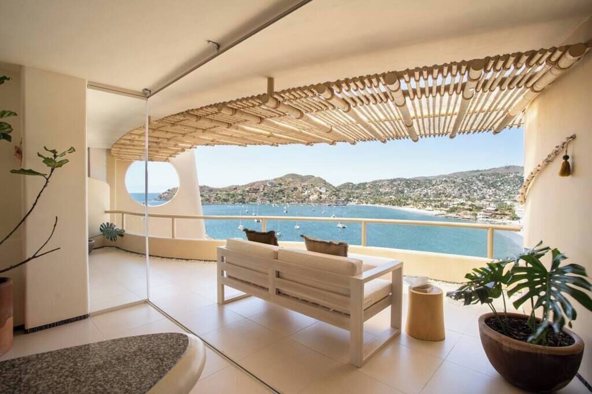 A balcony with a white bench, a potted plant, and a view of the sea and hills.