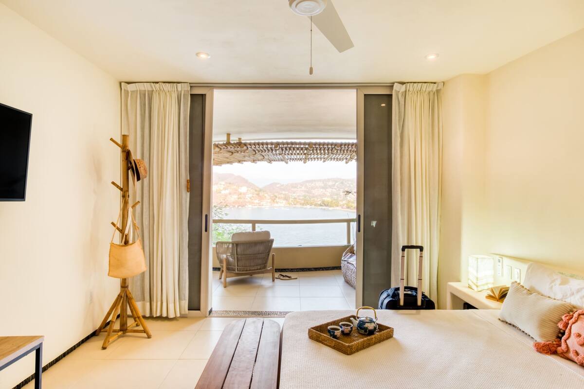 A hotel room with a bed, a table with a tray of tea, a chair, a suitcase, and a balcony with a view of the mountains.