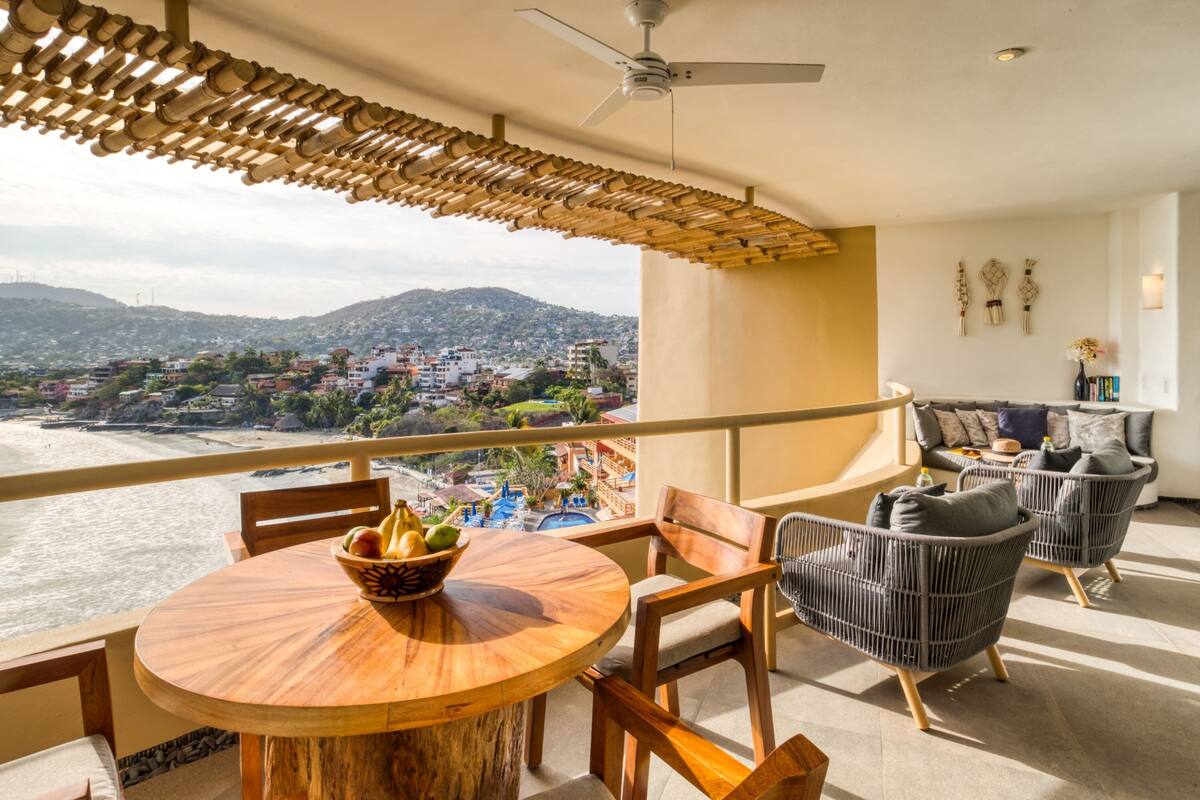 A balcony with a wooden table and chairs, a view of the beach and city, and a ceiling fan.