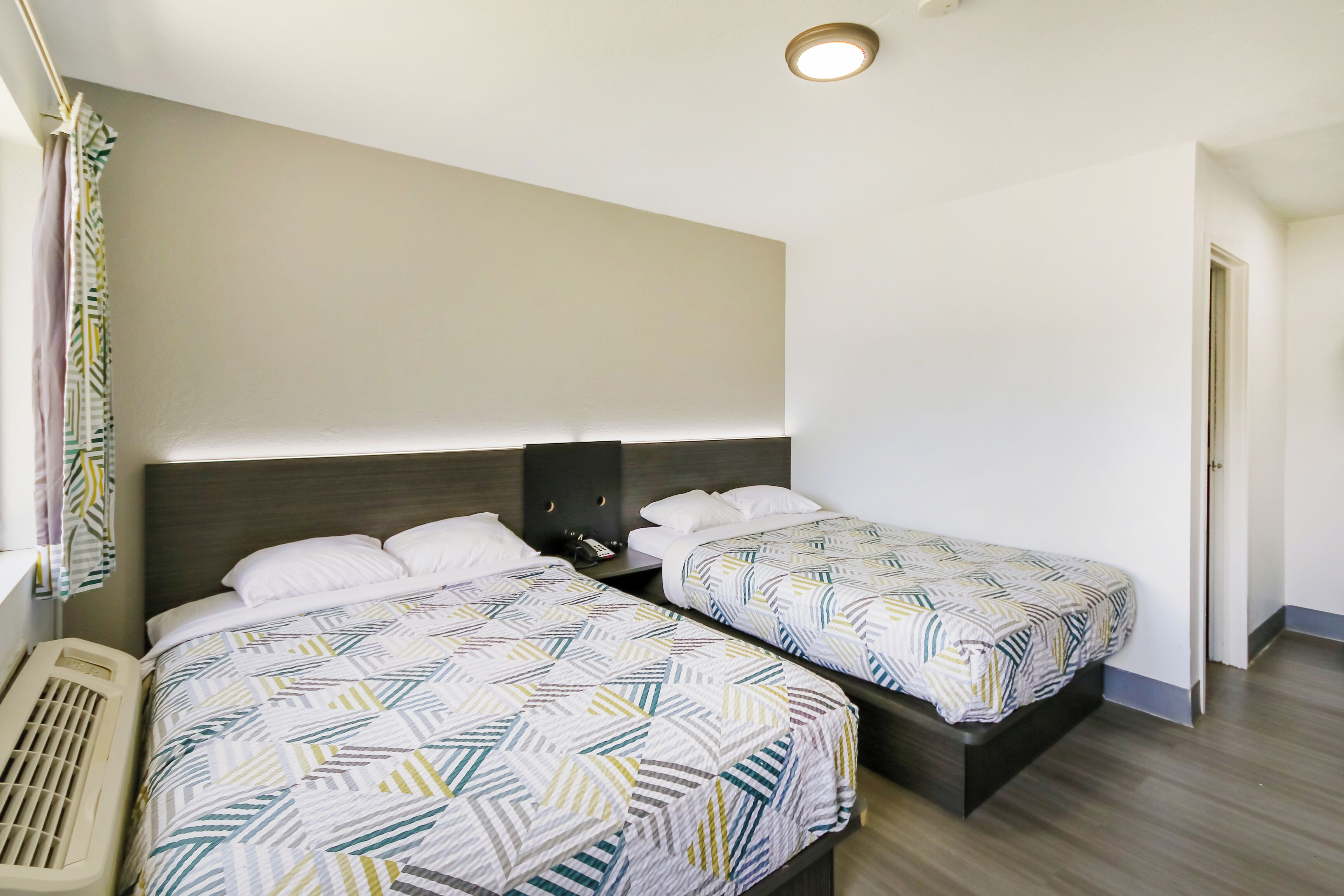 A hotel room with two beds, a ceiling light, a heater, and a door.