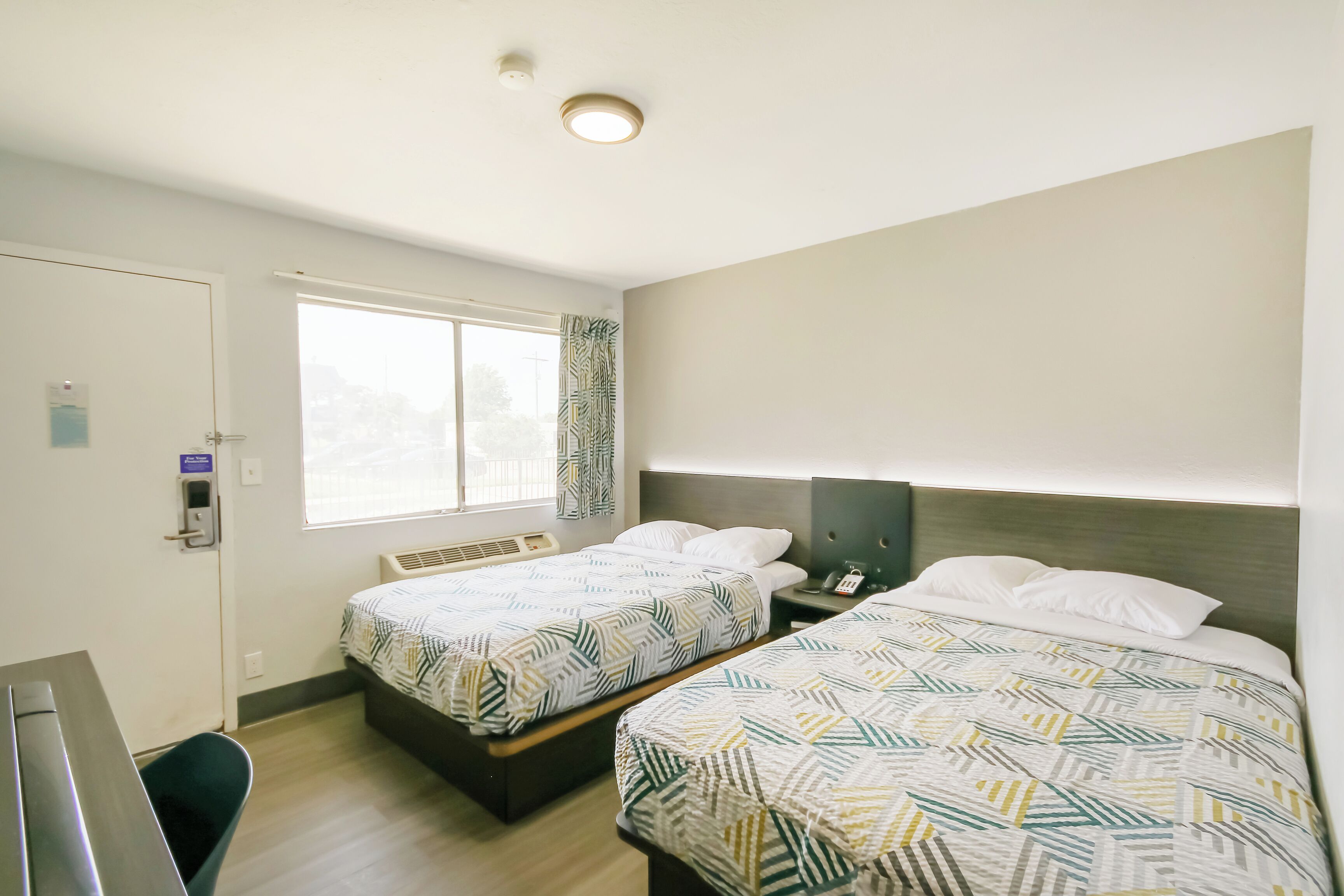 A hotel room with two beds, a desk, a chair, a window with curtains, and a ceiling light.