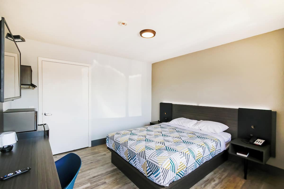 A hotel room with a bed, bedside table, nightstand, desk, chair, and a door.