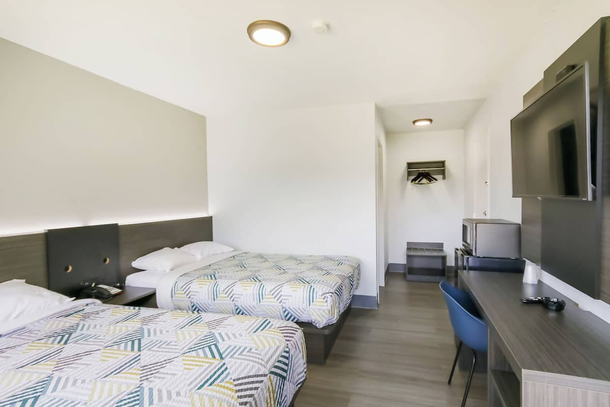 A hotel room with two beds, a desk, a chair, a TV, and a small kitchenette.