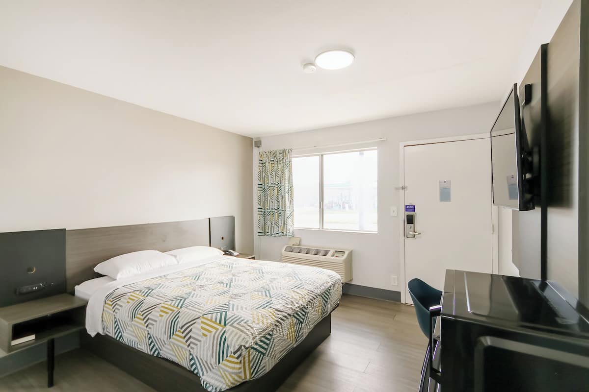 A modern hotel room with a bed, bedside table, desk, chair, TV, window with curtains, and an air conditioning unit.