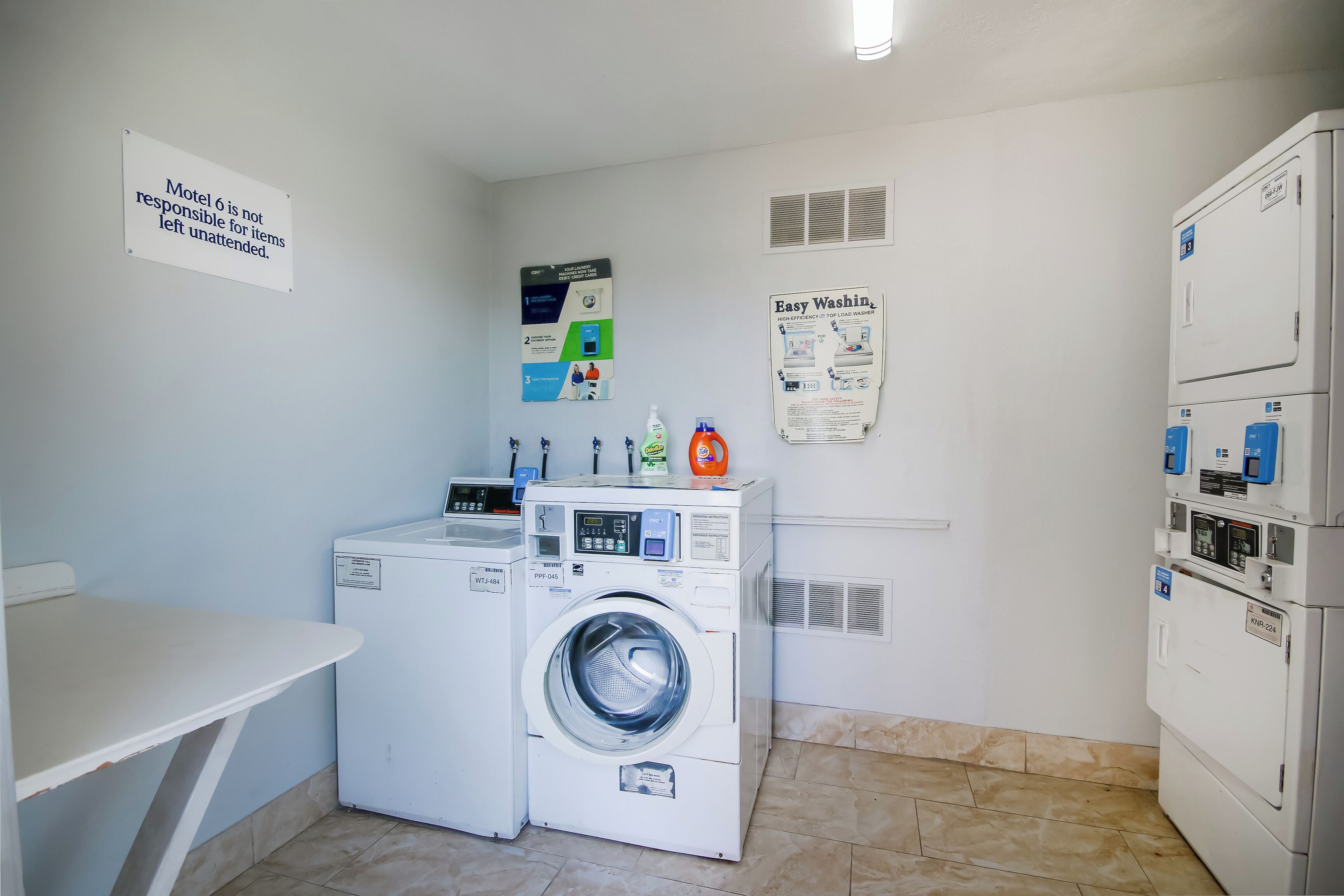 A laundry room with a washing machine, a dryer, and a table. There are signs on the wall, including one that says 'Motel is not responsible for items left unattended.'