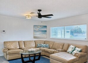 A living room with a beige sectional sofa, a glass coffee table, a ceiling fan, a beach-themed painting, and a window with blinds.