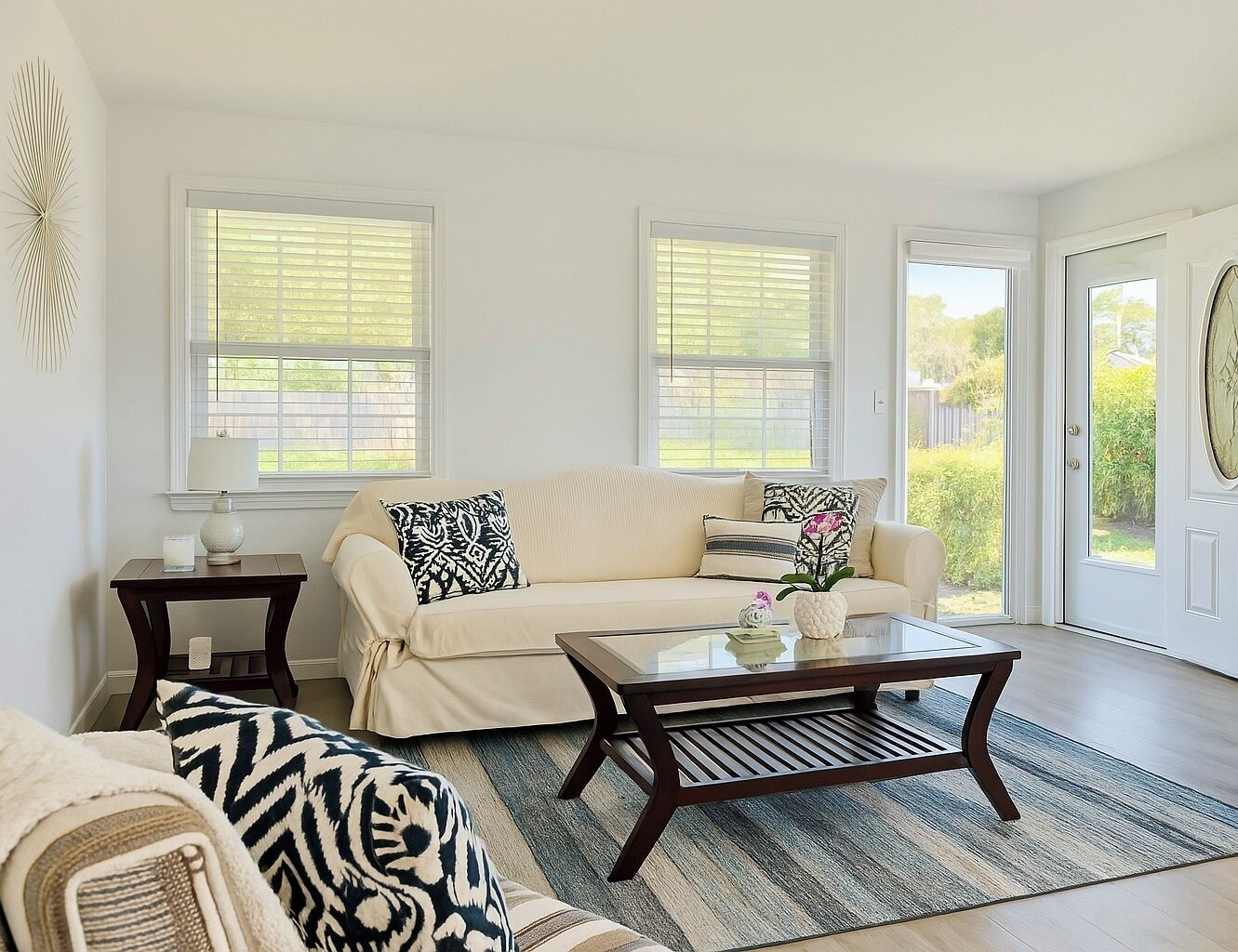 A living room with a beige sofa, a coffee table, and a patterned rug. There are two windows with blinds, a door, and a wall-mounted light fixture.