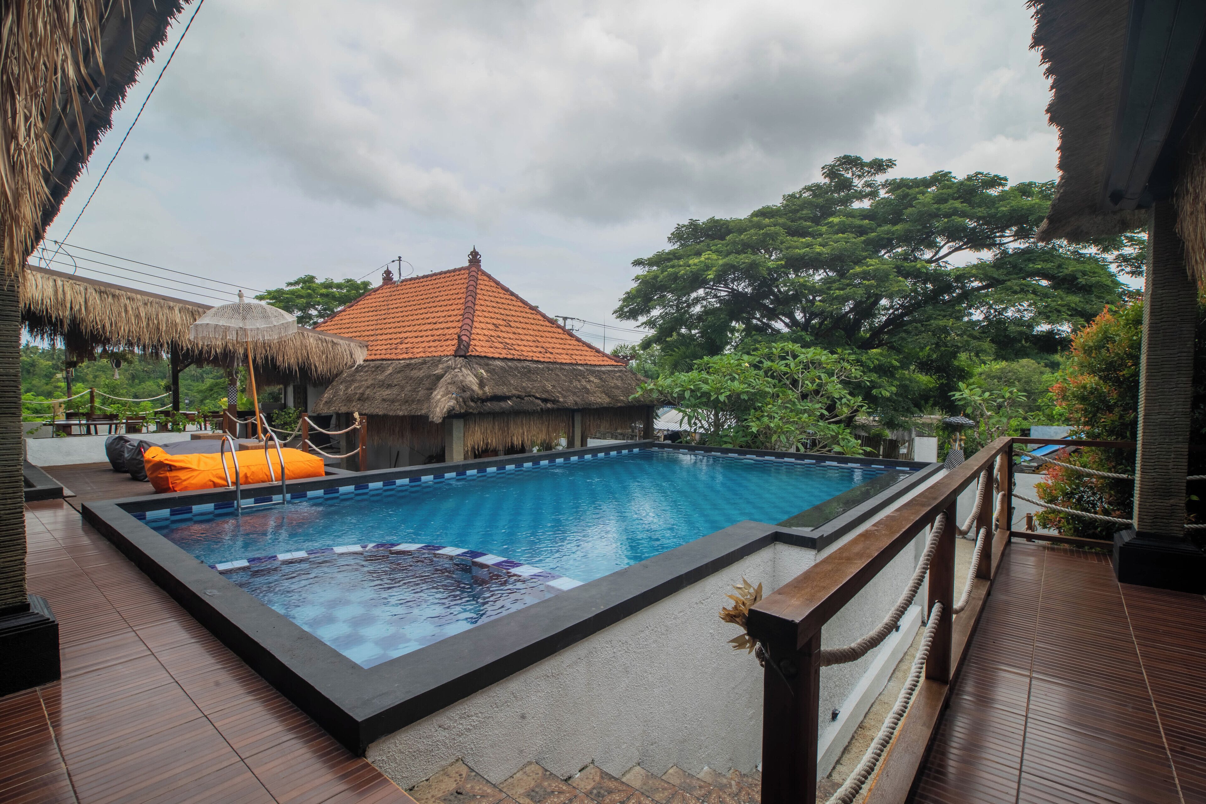 A swimming pool with a wooden deck, a thatched-roof structure, and lush greenery in the background.