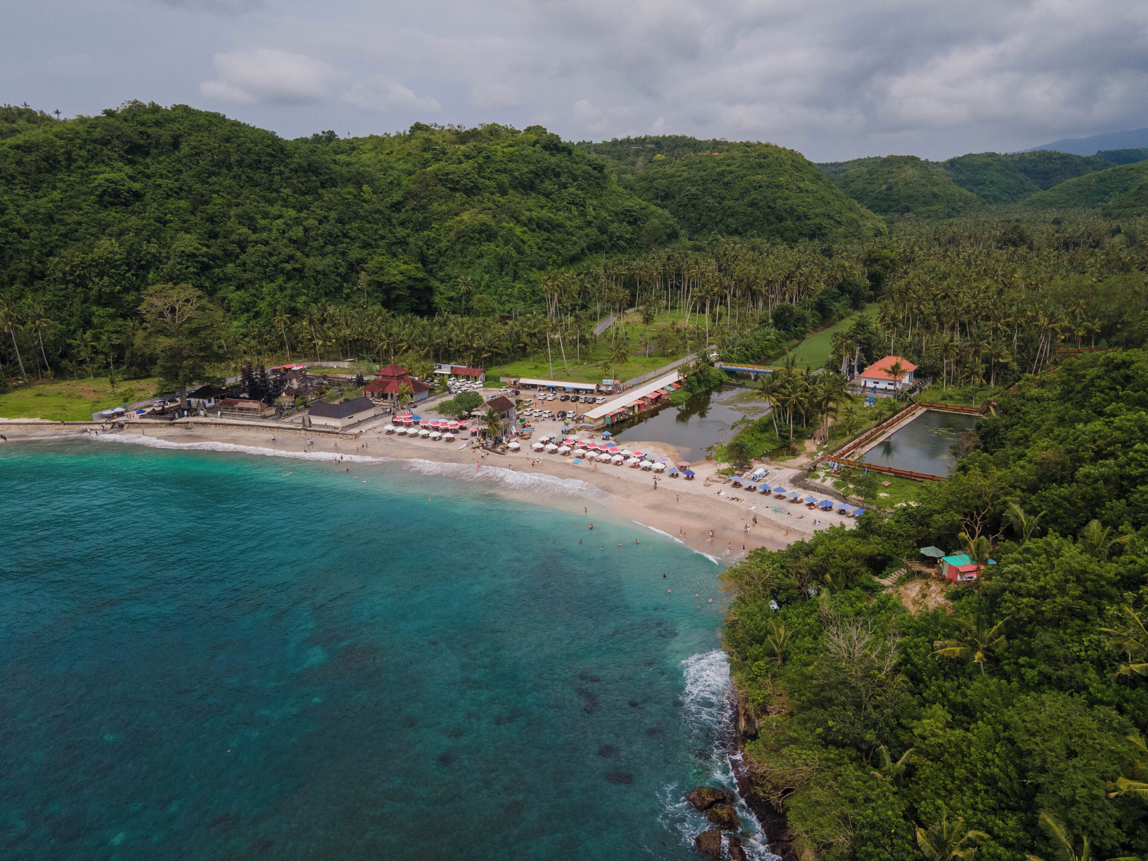 An aerial view of a coastal resort with a beach, clear turquoise waters, and lush green hills in the background.