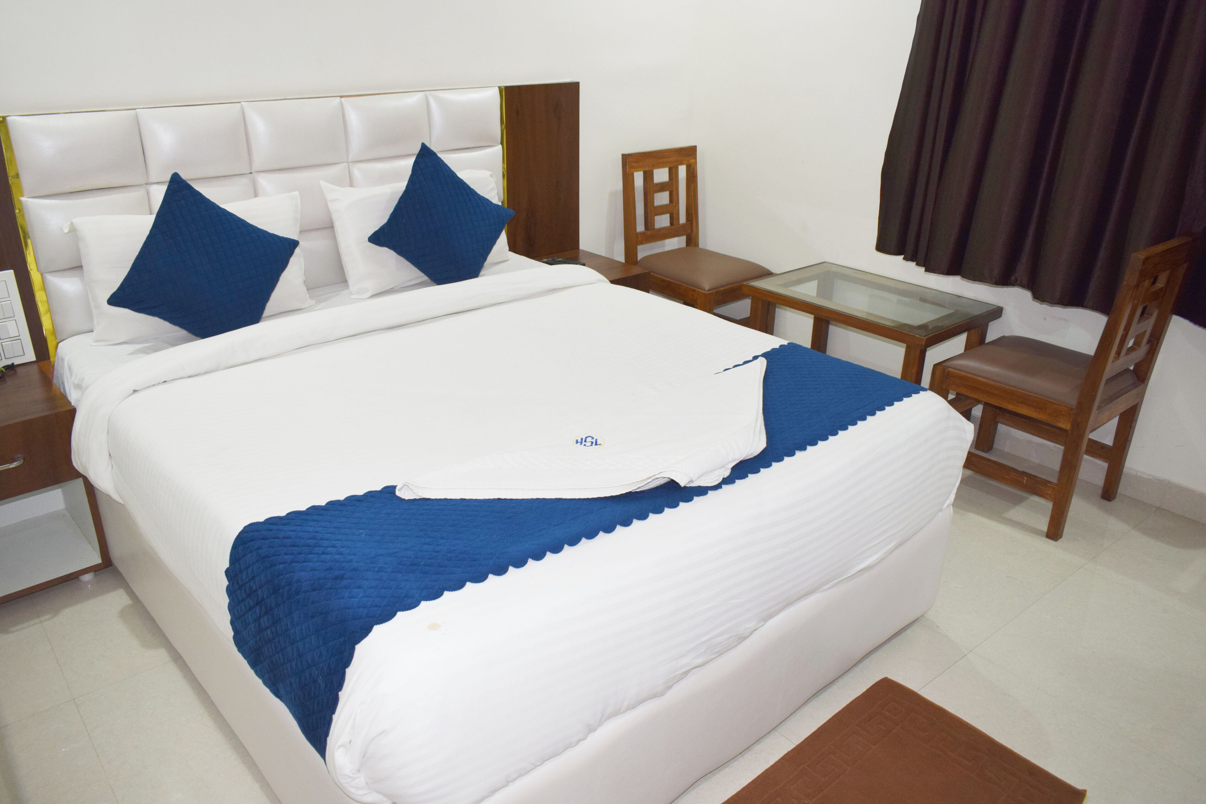 A neatly made bed with white and blue bedding, a wooden chair, a small glass table, and a brown rug.