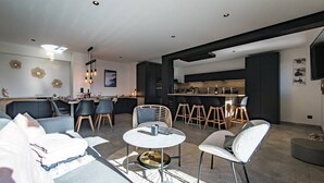 A modern living room with a dining area, a kitchen, and a bar. There are multiple seating options including a sofa, chairs, and a round table. - Sylodges 202 - Spacious apartment for 10/12 people (LA TOUSSUIRE)