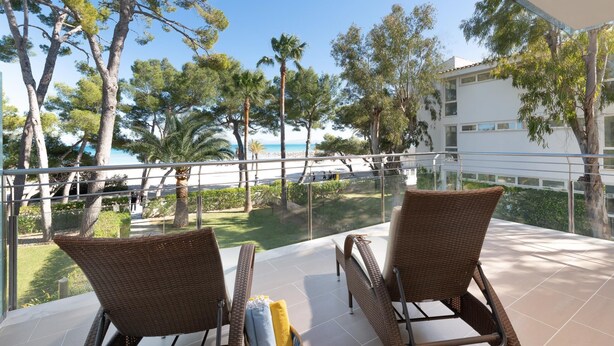 Un balcón con sillas de mimbre y una mesa, con vista a una playa y un edificio.