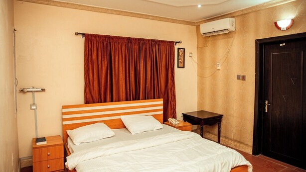 A hotel room with a bed, a nightstand, a bedside table, a wall-mounted air conditioner, and a door. - The CEO hotel and suite  (Ibadan)