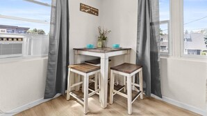 A corner room with a white table and two wooden stools, a small potted plant, and a window with grey curtains.