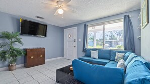 A living room with a blue sofa, a television mounted on the wall, a ceiling fan, and a window with curtains. - Waterfront Angler’s Paradise, 3BR/2.5BA w/ Dock, Boat Ramp, Kayaks, HorseShoes, CornHole, Ping Pong (New Port Richey)