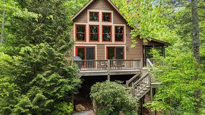 Une cabane dotée d’une terrasse et de nombreuses fenêtres, entourée d’une forêt dense.