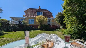 Ein Tisch mit Essensanordnung, darunter ein Fischgericht, Brot und ein Getränk, mit einem Haus im Hintergrund.