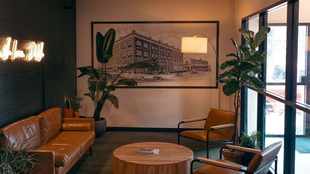 Een hal met een bruine leren bank, een ronde houten salontafel en twee metalen stoelen. Aan de muur hangt een groot schilderij van een stadsgezicht, een neonbord en staat een grote potplant.
