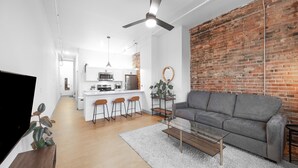 A modern living space with a kitchen, a grey sofa, and a brick accent wall. - Craft District Apt w/ Gym & Coffee Shop 225 (Duluth)