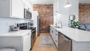 A modern kitchen with white cabinetry, stainless steel appliances, a brick accent wall, and pendant lighting. - Craft District Apt w/ Gym & Coffee Shop 225 (Duluth)