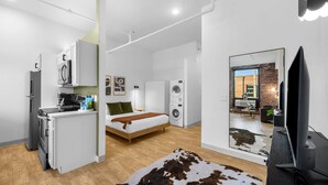 Un appartement moderne comprenant une cuisine, un lit, un lave-linge et un grand miroir qui reflète un espace de vie avec un téléviseur et un mur en briques.