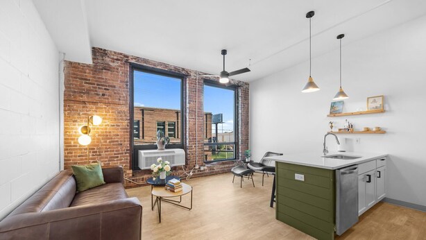 Un appartement moderne avec un mur en briques, une grande fenêtre, une cuisine dotée d’un plan de travail vert et un coin salon avec un canapé et une table basse.