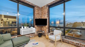 A modern living room with a large TV, comfortable seating, and a view of a parking lot. - Craft District Apt w/ Gym & Coffee Shop 207 (Duluth)