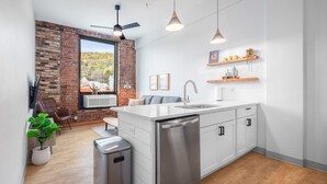 A modern kitchen with white cabinets, a stainless steel dishwasher, and a brick wall. There is a window with a view of trees and a ceiling fan.
