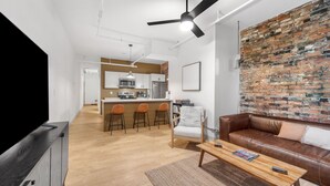 A modern living room with a brown leather sofa, a wooden coffee table, and a TV on a black stand. The room features a brick wall, a ceiling fan, and a kitchen area with a bar stool.