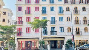 A multi-story building with a row of balconies, a ground-level entrance with a glass door, and a small outdoor seating area. - KIEN HOTEL (Phu Quoc)