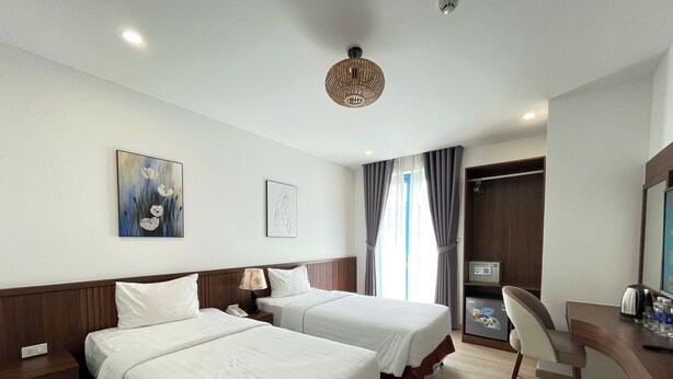 A hotel room with two beds, a desk with a chair, a minibar, and artwork on the walls. - KIEN HOTEL (Phu Quoc)