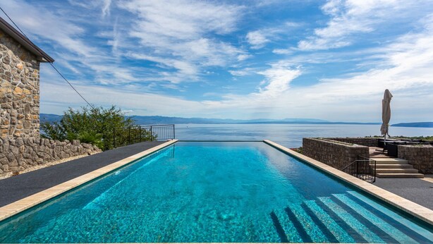 Una piscina rectangular con agua azul cristalina, rodeada de muros de piedra y un edificio de piedra, con vistas a una masa de agua y montañas distantes.