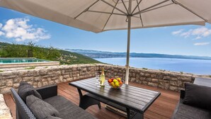 Un área de estar al aire libre con mesa, sillas y una sombrilla, con vista a un paisaje panorámico de una masa de agua y colinas lejanas.