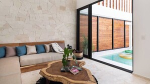 Un salon moderne avec un mur en pierre, un canapé en bois agrémenté de coussins, une table basse avec une plante, et une vue sur une piscine à travers de grandes fenêtres.