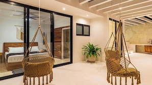 Une chambre d’hôtel moderne avec un grand lit, deux fauteuils suspendus, une plante en pot et une cloison en verre.