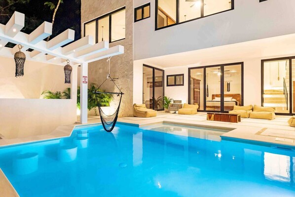 A modern indoor pool with a hammock, seating area, and large windows.