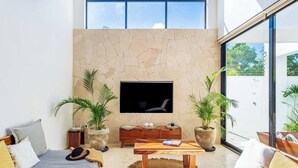 A modern living room with a stone wall, a flat-screen TV, two potted plants, a wooden coffee table, and a sofa with cushions. - New Spacious Villa with Rooftop dipping pool (Tulum)