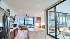 A modern kitchen with a central island, large windows, and a sliding glass door leading to an outdoor seating area. - New Spacious Villa with Rooftop dipping pool (Tulum)