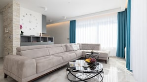 A modern living room with a beige sectional sofa, a glass coffee table with fruits, a magazine, and a cup of coffee. There is a bookshelf with decorative items and a wall clock.