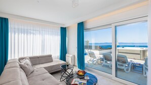 A modern living room with a sofa, a glass coffee table, and a view of the sea through large windows.