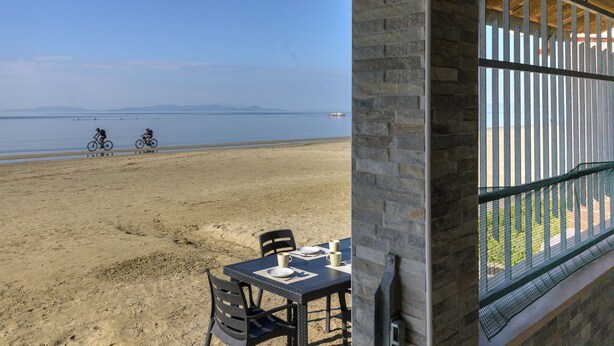 Uitzicht op het strand vanaf een overdekt terras, met een tafel en stoelen, met zicht op het strand en een bergketen in de verte.