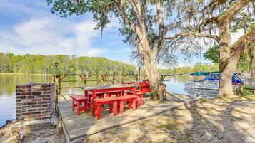 Une terrasse en bois avec des bancs rouges et des tables de pique-nique, située au bord d’un lac, entourée d’arbres et d’un quai.
