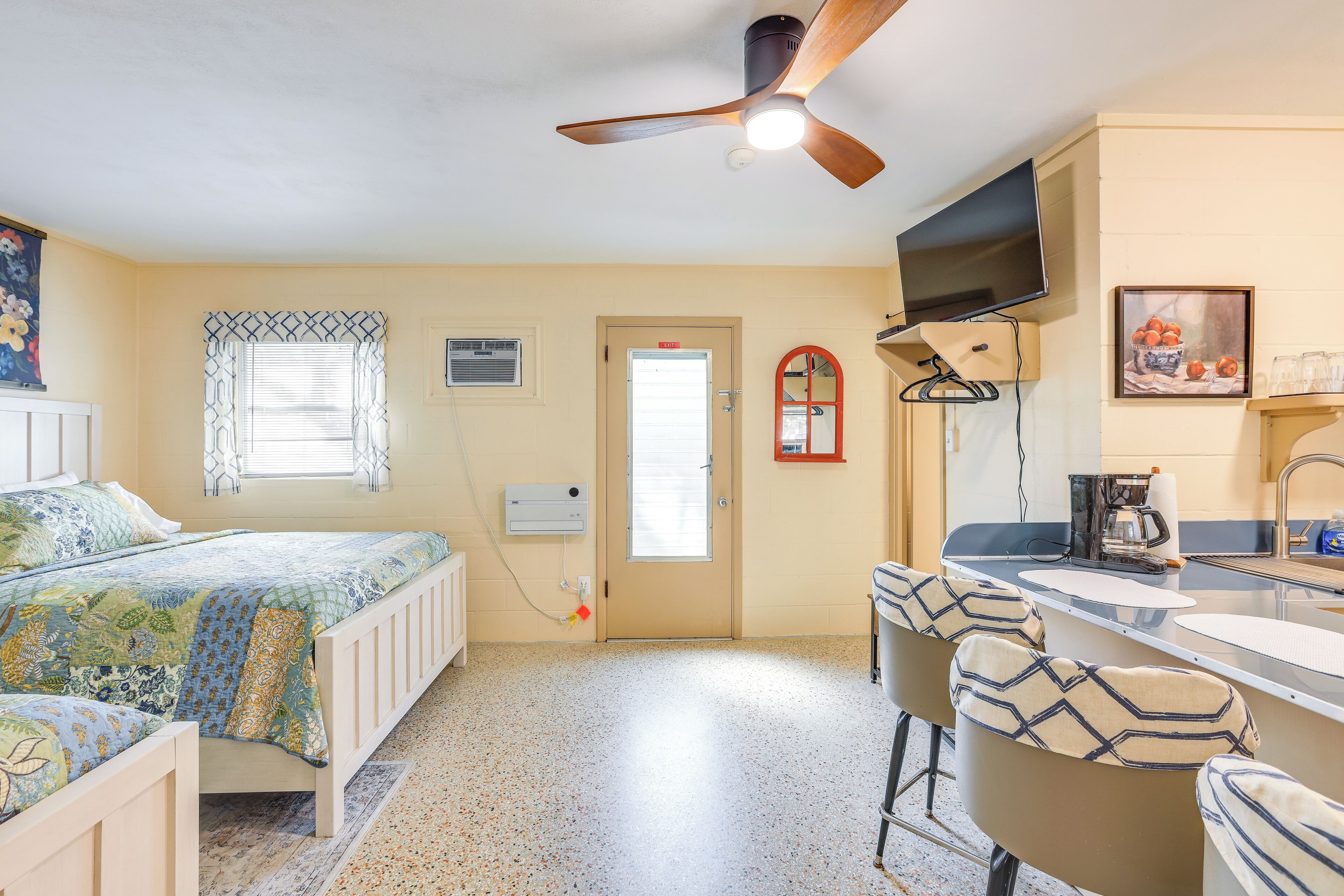 Une chambre compacte comprenant un lit, un ventilateur de plafond, une fenêtre avec des rideaux à motifs, une porte et un coin cuisine avec un évier et des tabourets de bar.