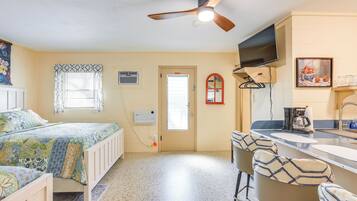 Une chambre compacte comprenant un lit, un ventilateur de plafond, une fenêtre avec des rideaux à motifs, une porte et un coin cuisine avec un évier et des tabourets de bar.