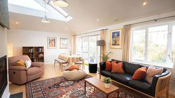 A living room with a sofa, a coffee table, a bean bag, a bookshelf, a dining table, and a large window with curtains.