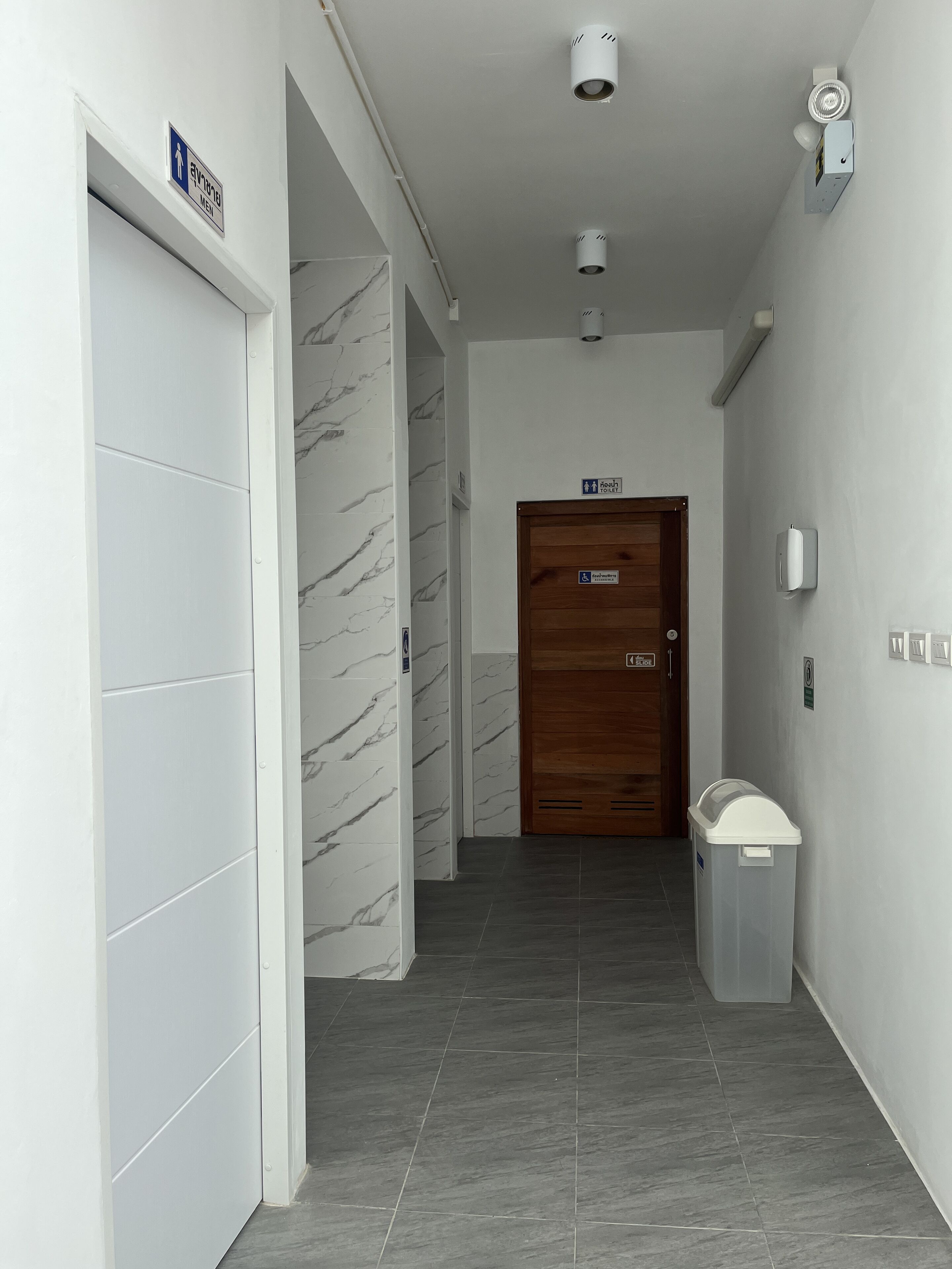 A hallway with a marble wall, a wooden door labeled "Men's Toilet," and a trash bin.
