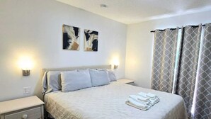 Ein ordentlich gemachtes Bett mit weißer Decke und Kissen, ein Nachttisch mit Lampe und ein an der Wand hängendes Kunstwerk.